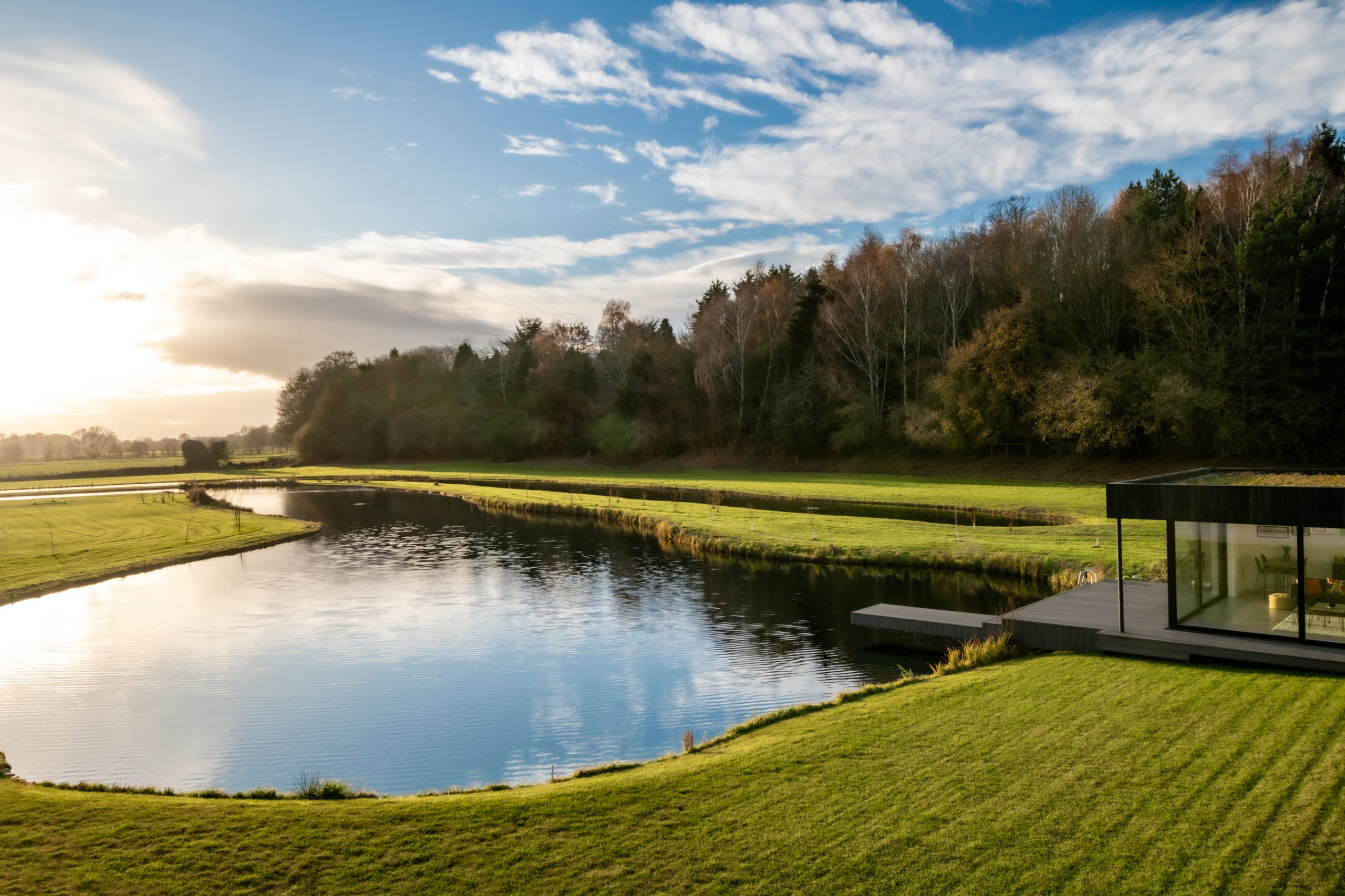 Modern glass pavilion with wooden deck beside a serene pond surrounded by manicured lawns and woodland at golden hour